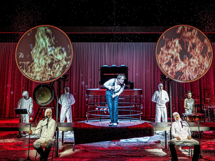 Eine Theateraufführung mit Schauspielern in weißen Kostümen vor einem roten Bühnenhintergrund. Zwei große, beleuchtete Scheiben sind sichtbar.