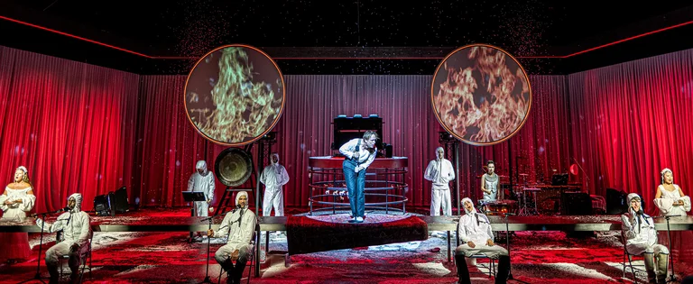 Eine Theateraufführung mit Schauspielern in weißen Kostümen vor einem roten Bühnenhintergrund. Zwei große, beleuchtete Scheiben sind sichtbar.