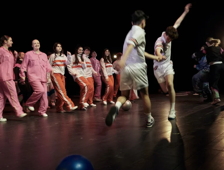 Eine Gruppe von Menschen auf einer Bühne. Zwei Personen in weißen Outfits tanzen in der Mitte, Zuschauer im Hintergrund.