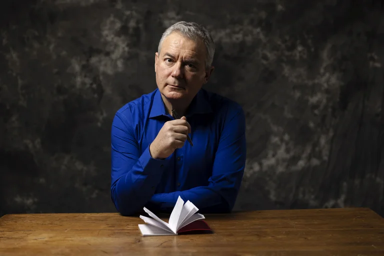 Ilija Trojanow in einem blauen Hemd sitzt an einem Tisch vor einem aufgeschlagenen Buch und denkt nach.