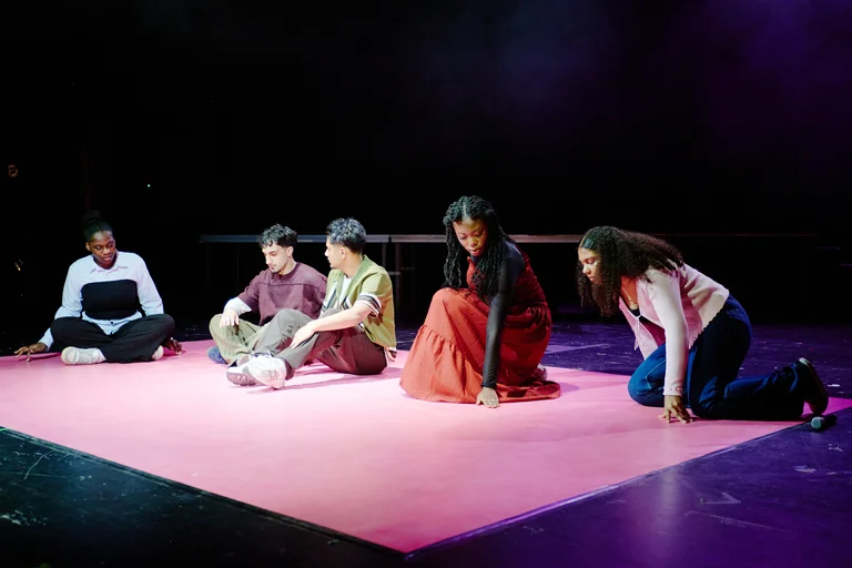 Fünf Personen sitzen auf einer pinken Bühne im Theater. Sie tragen legere Kleidung und sind in Gespräch oder Probe vertieft.