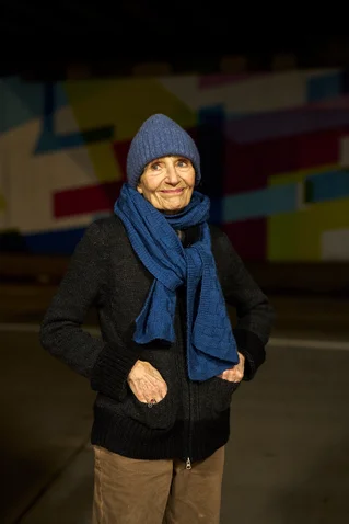 Eine Frau mit blauem Schal und Mütze steht vor einer farbenfrohen Wand. Sie trägt braune Hose und dunkle Jacke.