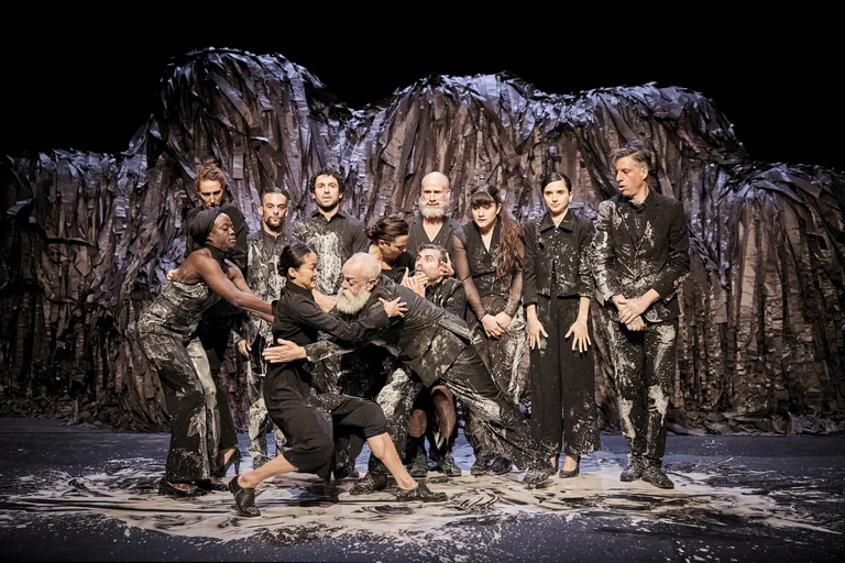 Ensemble von Tänzern in schwarz-silberner Kleidung vor einer dramatischen, bergartigen Kulisse. Intensive emotionale Ausdruckskraft.