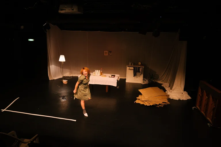 Eine Frau tanzt auf einer dunklen Theaterbühne. Die Bühne ist spärlich mit einem Tisch, Lampe und Dekorationen eingerichtet.
