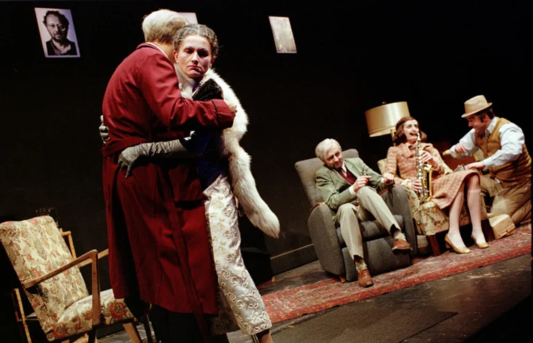 Eine Bühnenaufnahme einer Theateraufführung mit fünf Schauspielern in Vintage-Kleidung. Zwei umarmen sich, während drei auf Stühlen sitzen.