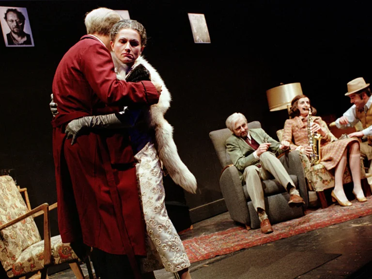 Eine Bühnenaufnahme einer Theateraufführung mit fünf Schauspielern in Vintage-Kleidung. Zwei umarmen sich, während drei auf Stühlen sitzen.