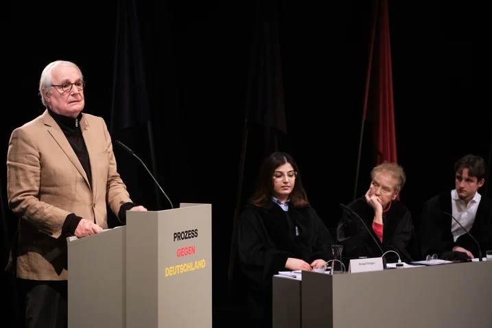 Eine Person spricht am Rednerpult bei einer Podiumsdiskussion. Drei Personen sitzen am Tisch und hören aufmerksam zu.