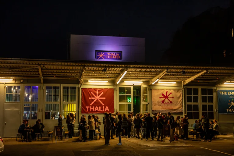 Night scene outside a building with lit windows and two large banners, people gathered beneath them, some seated, others standing.