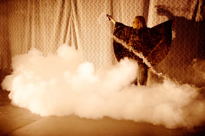 Eine Person in einem dramatischen Umhang steht auf einer Bühne mit dichtem, weißen Nebel. Die Beleuchtung erzeugt einen mystischen Effekt.