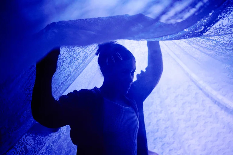 Eine Person hält im blauen Licht einen durchscheinenden Stoff über sich. Der Umriss ist im schattenhaften Kontrast sichtbar.