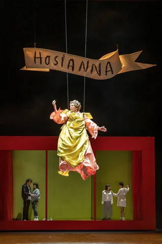 A stage performance featuring a suspended figure in a yellow costume, below a red frame. Banner reads "Hosianna."