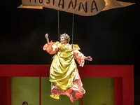 A stage performance featuring a suspended figure in a yellow costume, below a red frame. Banner reads "Hosianna."