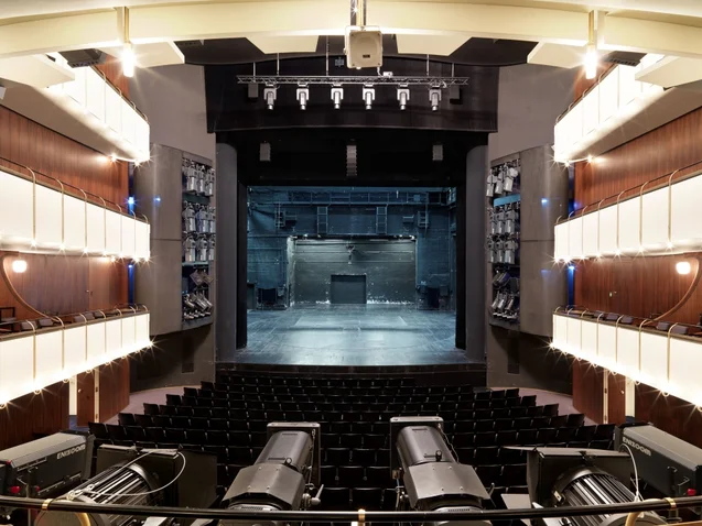 Ein leerer, moderner Theaterraum mit einer Bühne, umgeben von Sitzplätzen. Die Beleuchtung ist warm, der Vorführraum wartet auf Publikum.