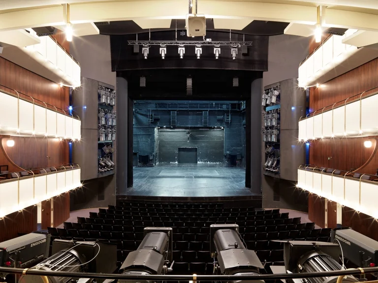 A modern theater auditorium with an empty stage, surrounded by seating. Warm lighting creates an inviting atmosphere, awaiting an audience.