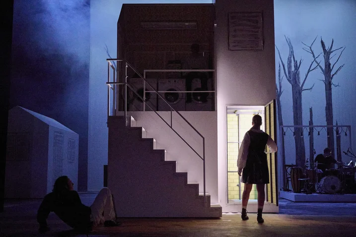 A stage set with a house featuring stairs. One person stands in the doorway, another sits on the floor.