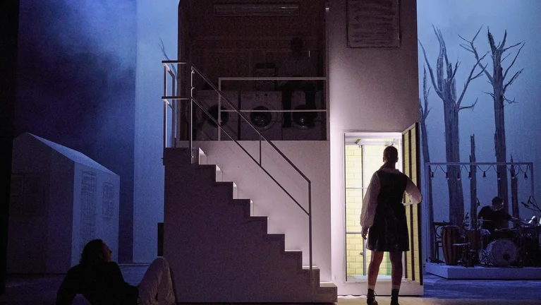 Eine Bühnenkulisse mit einem Haus mit Treppe. Eine Person steht in der Tür, während eine andere am Boden sitzt.