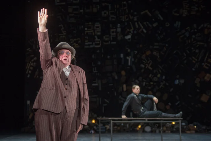An actor in a brown suit raises his hand on stage. Another actor sits in the background on a bench.