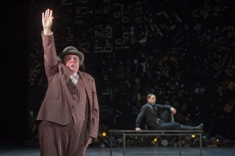 Ein Schauspieler in einem braunen Anzug hebt die Hand auf einer Bühne. Ein anderer Schauspieler sitzt im Hintergrund auf einer Bank.