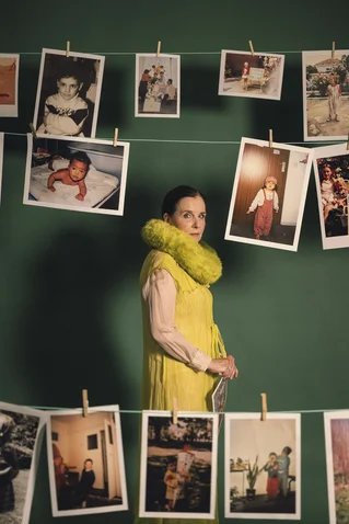 A woman in a yellow dress stands against a green backdrop, surrounded by hanging photos on lines. Vintage photo exhibition.