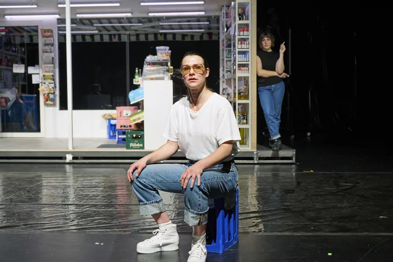 Ein Schauspieler sitzt auf einem blauen Hocker vor einem stilisierten Laden. Der Hintergrund ist dunkel und minimalistisch gestaltet.