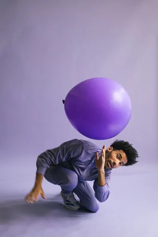 A man in a gray tracksuit balances a large purple balloon. He kneels on a light studio floor.