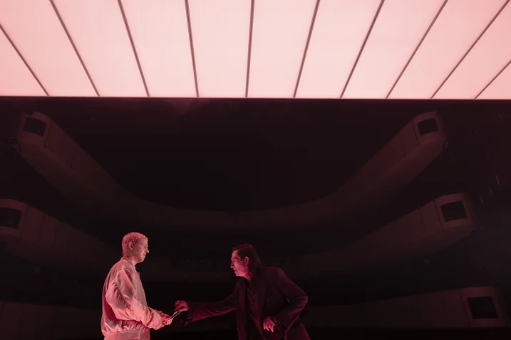 Two people on stage under a brightly lit ceiling, seemingly engaged in a dramatic or artistic performance.