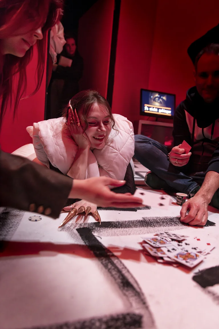In einem Raum mit rotem Licht spielen Personen mit Spielkarten auf einem Tisch. Eine Person stützt ihren Kopf auf die Hand.