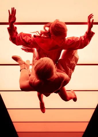 Two dancers in motion, illuminated by intense red light, creating a dramatic, floating illusion.