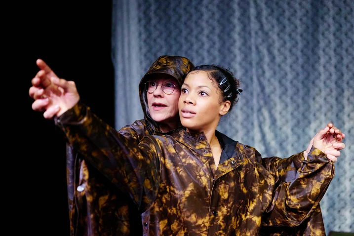 Zwei Personen in Kostümen auf einer Bühne, weisen dramatisch in eine Richtung. Der Hintergrund hat ein welliges Muster.