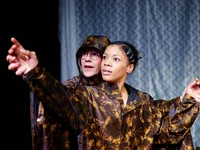 Two people in costumes on stage, pointing dramatically. The background features a wavy pattern.