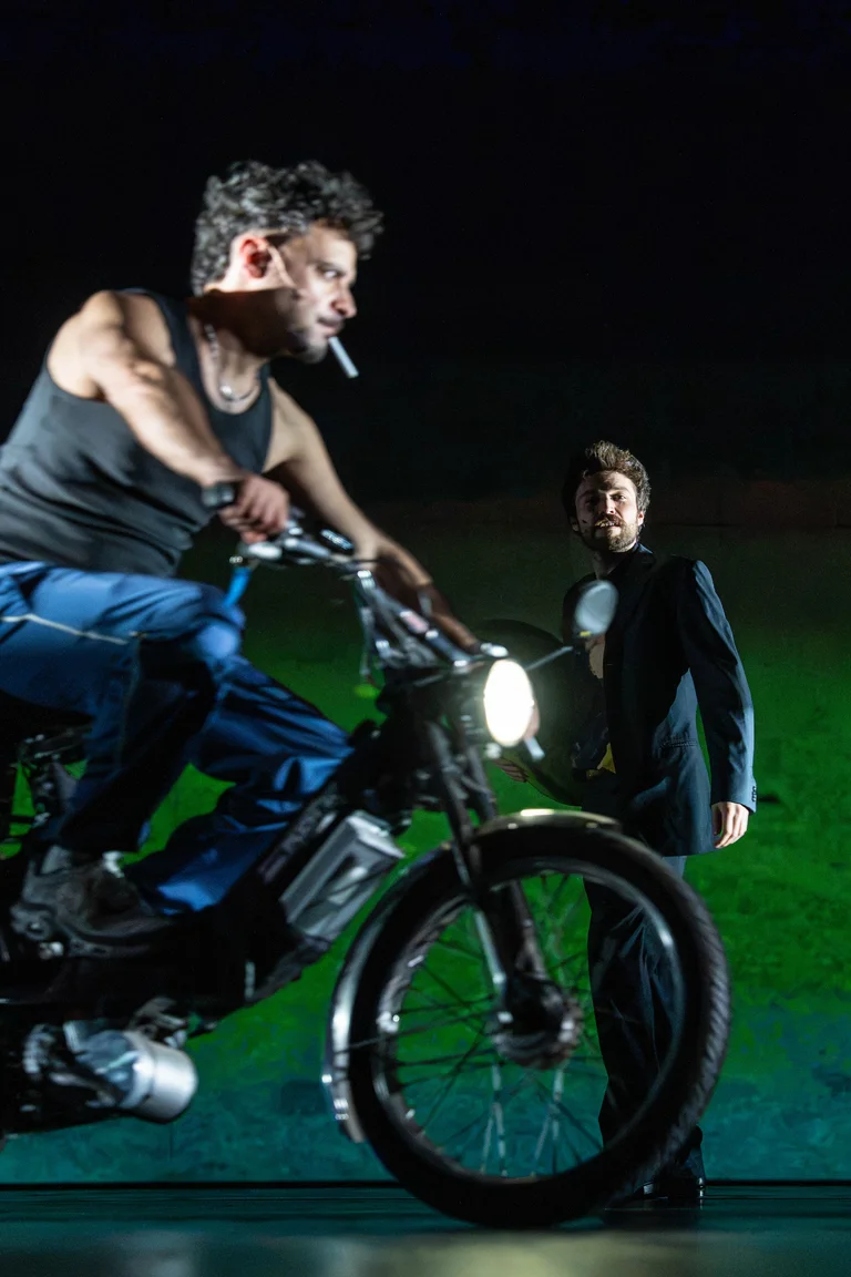 Ein Mann auf einem Motorrad fährt im Vordergrund, während ein anderer Mann im Anzug im Hintergrund steht und zusieht.