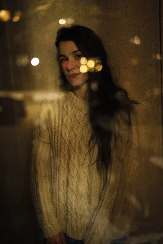 Eine Person in einem cremefarbenen Pullover steht hinter Glas, mit reflektierten Lichtern im Hintergrund.
