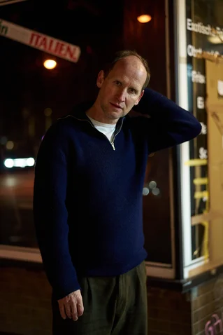 Ein Mann steht lässig in einem dunklen Pullover vor einem Geschäft bei Nacht. Neonschilder sind im Hintergrund.