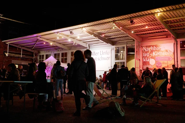Menschen versammeln sich nachts unter einem beleuchteten Pavillon. Einige sitzen auf Liegestühlen, andere stehen und unterhalten sich.