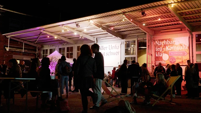 Menschen versammeln sich nachts unter einem beleuchteten Pavillon. Einige sitzen auf Liegestühlen, andere stehen und unterhalten sich.