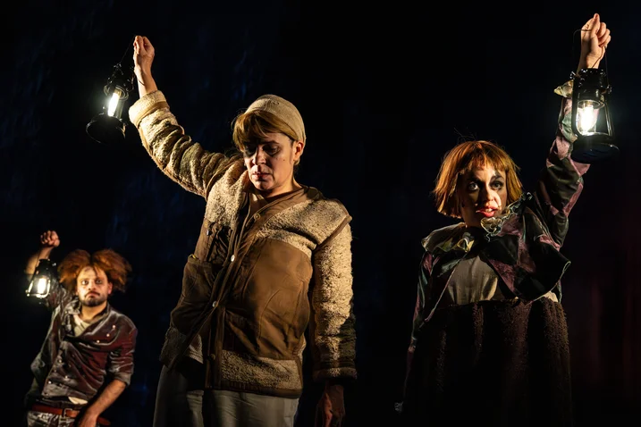 Three people in costumes hold lanterns high on a dark stage, as if depicting a scene from a play.
