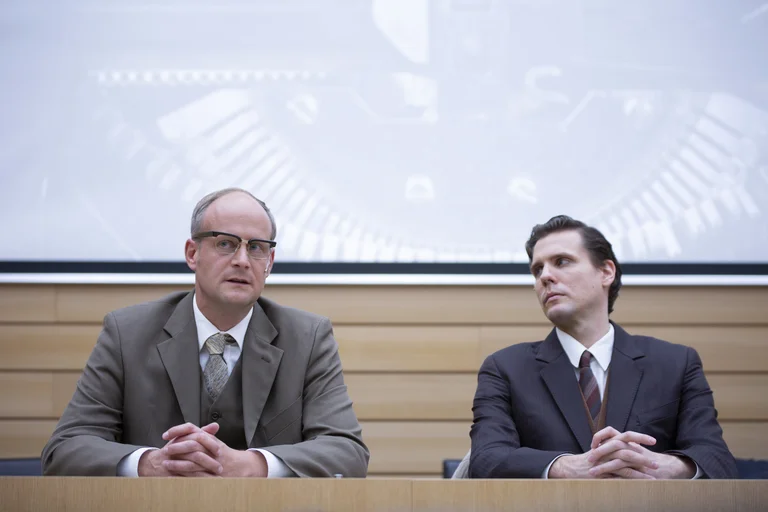 Zwei Männer in Anzügen sitzen an einem Konferenztisch vor einer Leinwand. Sie haben stille, nachdenkliche Ausdrücke.