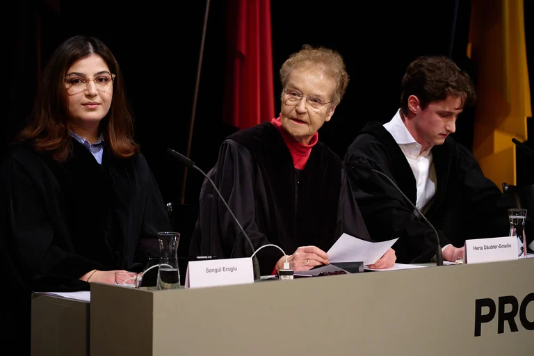 Drei Personen sitzen nebeneinander an einem Tisch mit Mikrofonen. Sie tragen dunkle Roben und halten Papiere in der Hand.