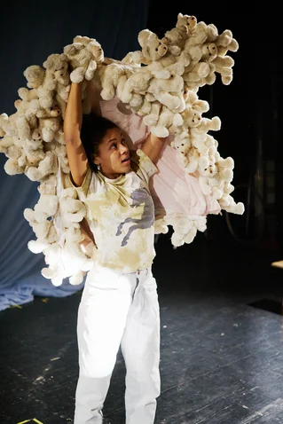 Eine Person hebt ein großes Bündel weißer Plüschtiere über den Kopf, stehend in einem dunklen Raum mit Bühnenbeleuchtung.