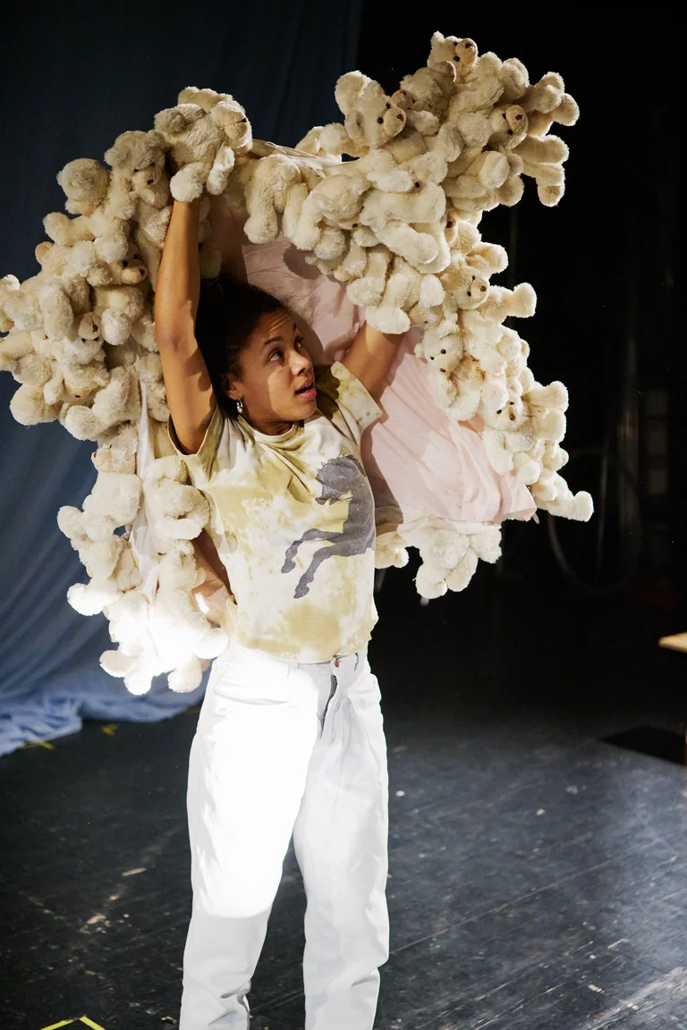 Eine Person hebt ein großes Bündel weißer Plüschtiere über den Kopf, stehend in einem dunklen Raum mit Bühnenbeleuchtung.