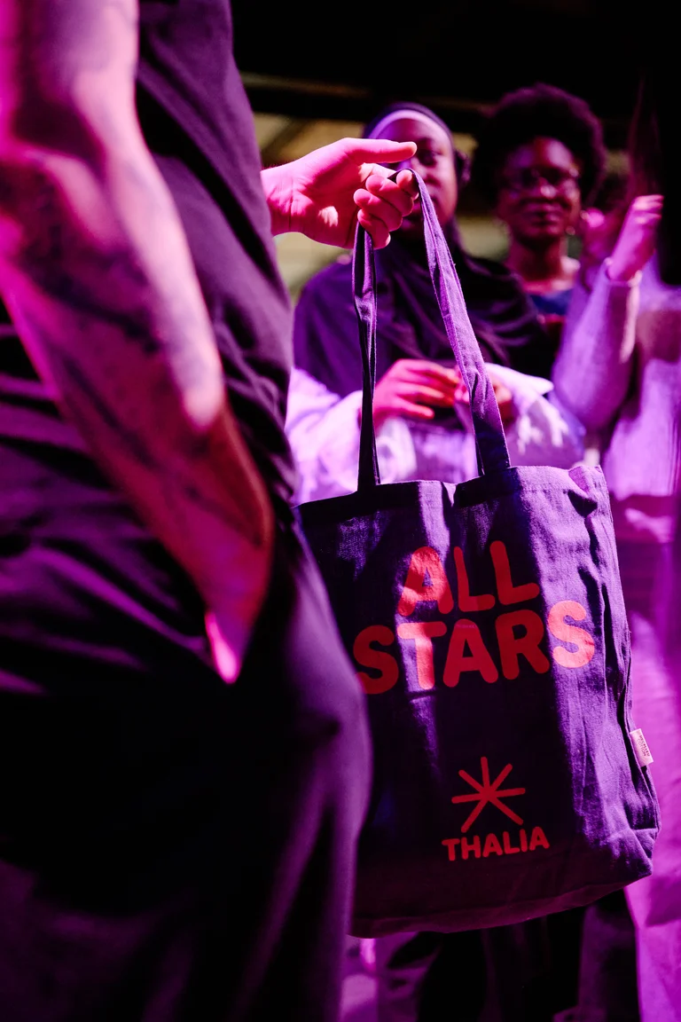 Eine Person hält eine Tasche mit der Aufschrift "All Stars". Im Hintergrund lächeln mehrere Menschen, warme Beleuchtung.