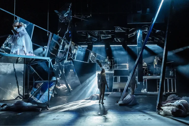 Ein düster beleuchtetes Theaterstück mit dramatischen Kulissen und Schauspielern. Die Bühne ist voller schräger, fragmentierter Strukturen und intensivem Licht.