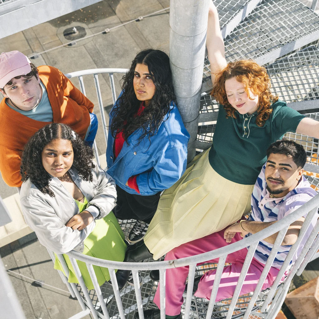 Fünf junge Erwachsene posieren auf einer Wendeltreppe im Freien. Sie tragen bunte Kleidung und blicken optimistisch in die Kamera.