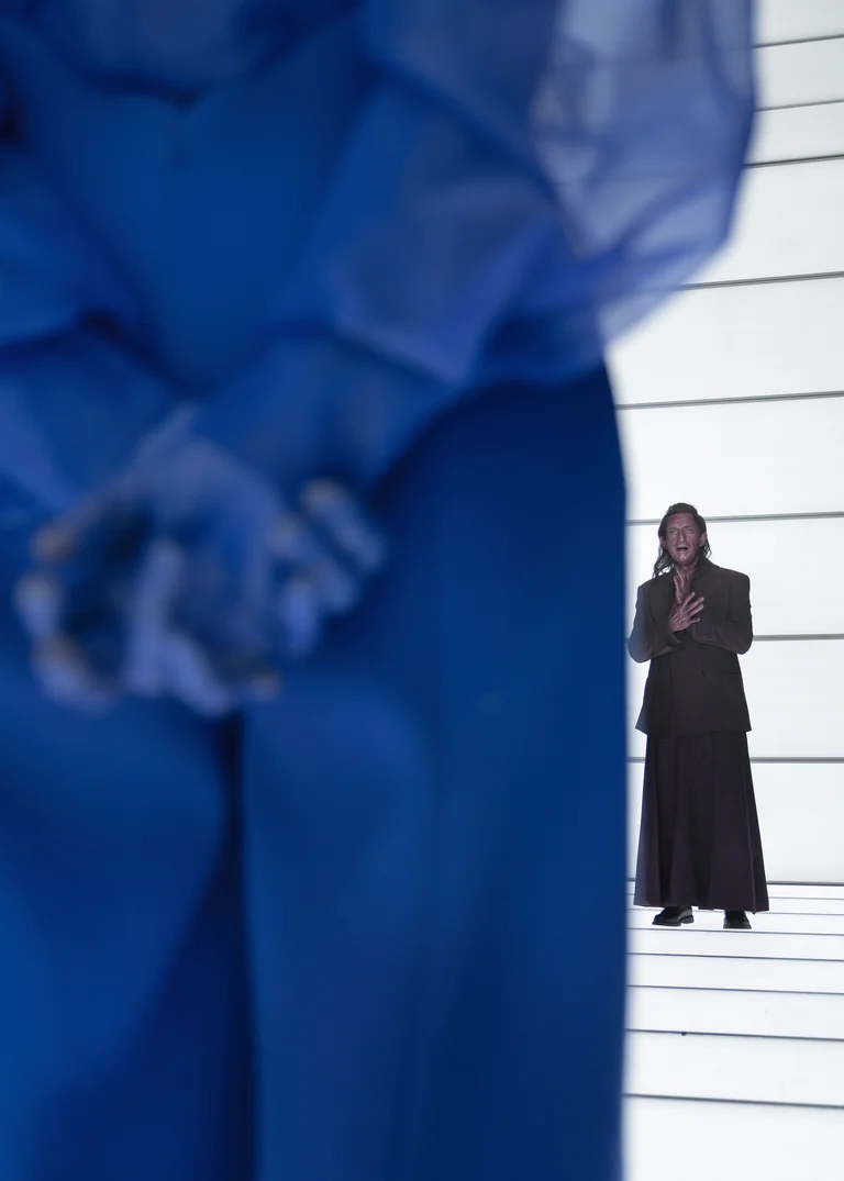 A person in a blue costume stands in the foreground, with another in black in the background on a striped stage.