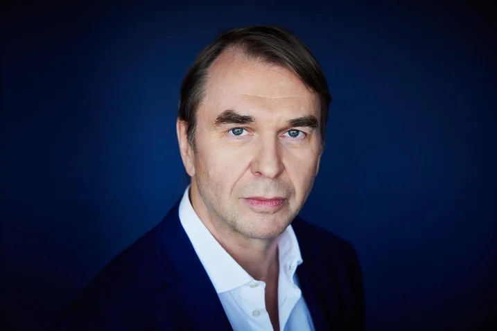 A man with dark hair wearing a white shirt and a dark blue suit against a blue background.