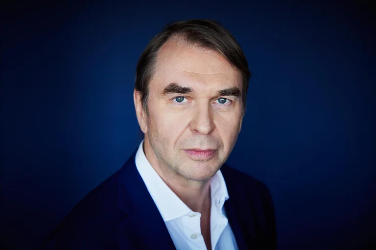 Ein Mann mit dunklem Haar trägt ein weißes Hemd und einen dunkelblauen Anzug vor einem blauen Hintergrund.