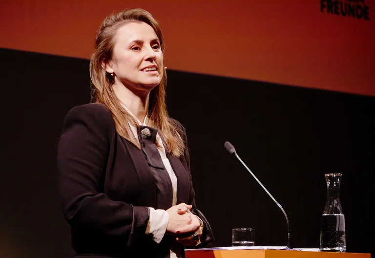 Eine Frau in einem dunklen Blazer steht an einem Rednerpult und spricht vor einem orangefarbenen Hintergrund.