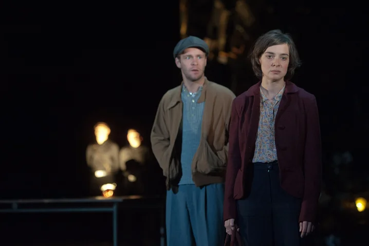 Two actors stand on a dark stage. The man wears a cap, the woman looks serious.