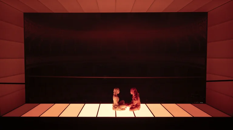 Two people sit on a lit stage with a minimalist background. The atmosphere appears dramatic and mysterious.