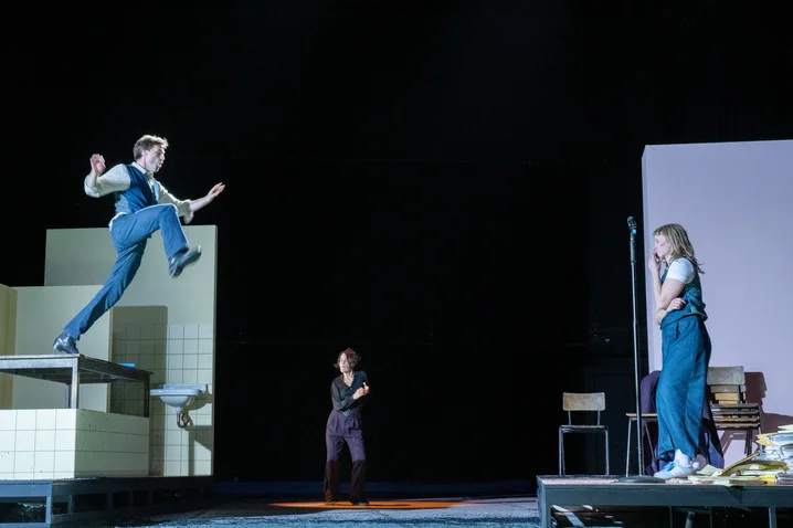 Three people on a stage: One person jumps off a platform, while two others watch. Dramatic lighting.